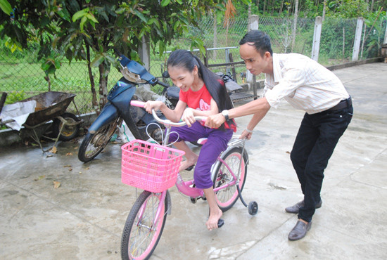 Tình nguyện viên Hội Chữ thập đỏ Tiên Thọ giúp đỡ em Hồ Thị Hồng Linh di chuyển xe đạp quanh sân nhà. Ảnh: N.HƯNG