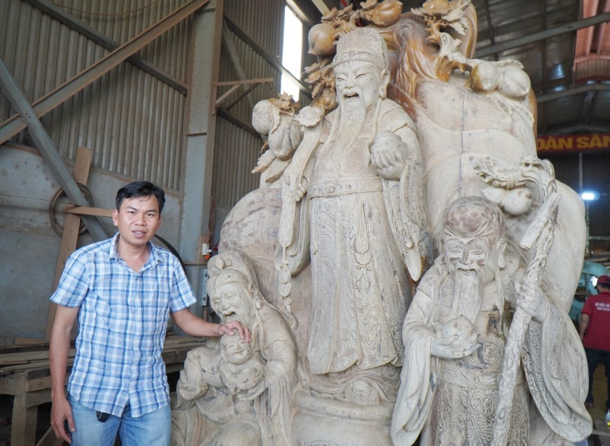 Trần Hữu Nhựt bên pho tượng Phúc Lộc Thọ bằng gỗ mun hoa đang hoàn thiện tại nhà xưởng Công ty Ngọc Trị. Ảnh: T.BÌNH