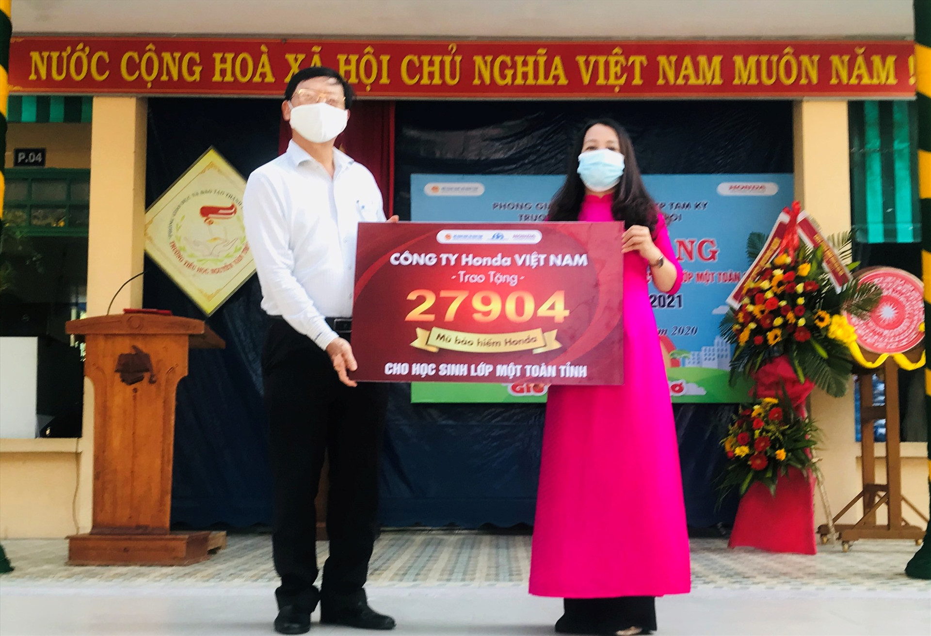 Hiệu trưởng Trường Tiểu học Nguyễn Văn Trỗi, Tam Kỳ (phải) tiếp nhận bảng tượng trưng 27.904 mũ bảo hiểm của Công ty Honda Việt Nam từ Ban An toàn giao thông tỉnh trao tặng từ cho học sinh lớp 1 năm học 2020-2021 tại Quảng Nam.