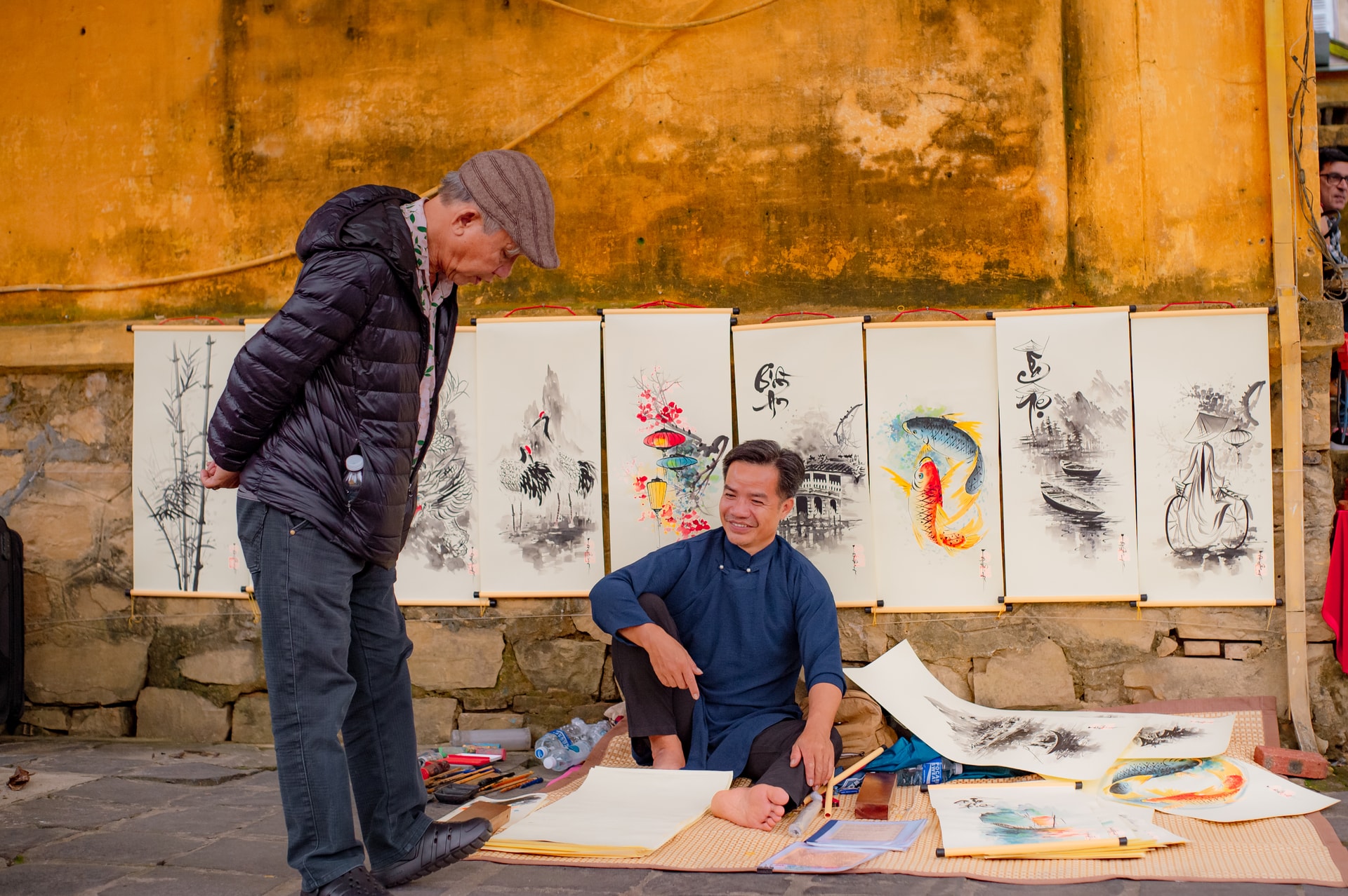 Un coin de la vieille ville de Hoi An, avec des peintures accrochées le long des murs jaunes ; l'artiste est assis et discute avec les visiteurs qui admirent les tableaux, créant une atmosphère chaleureuse et quotidienne.